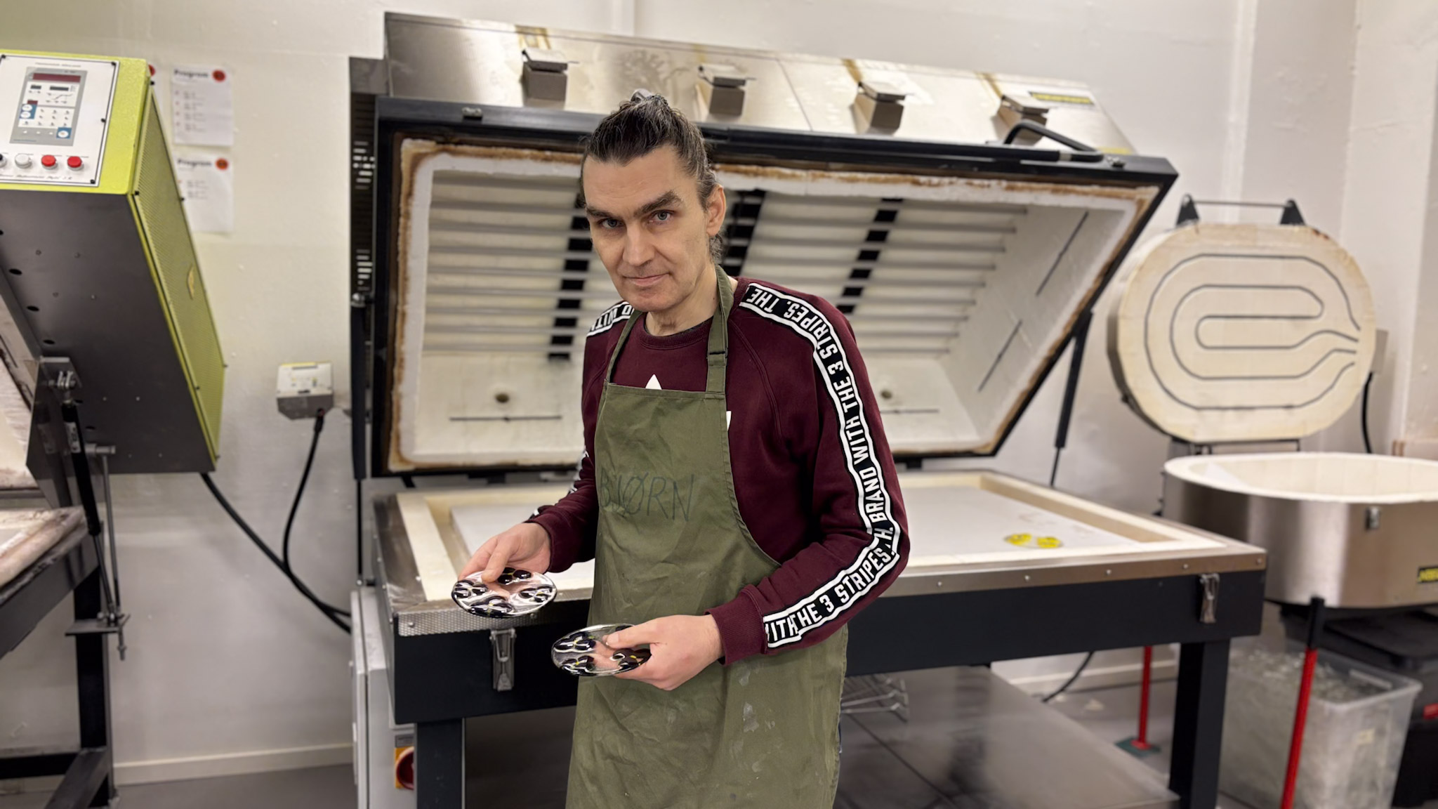 Bj&oslash;rn holder opp to glassprodukter han har laget. Han st&aring;r foran verkstedets ovn.