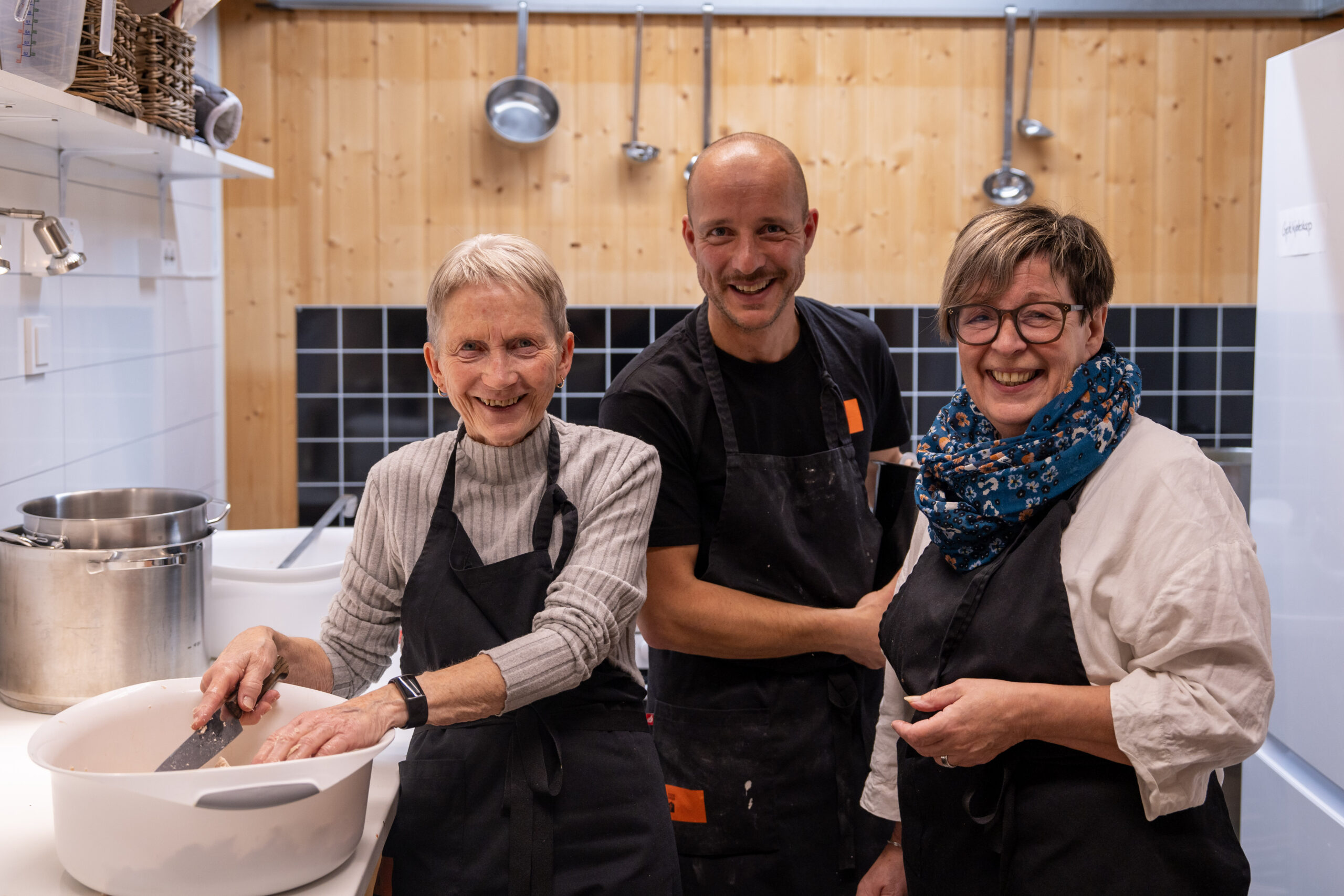 To kvinner og en mann som lager mat og smiler mot kamera.