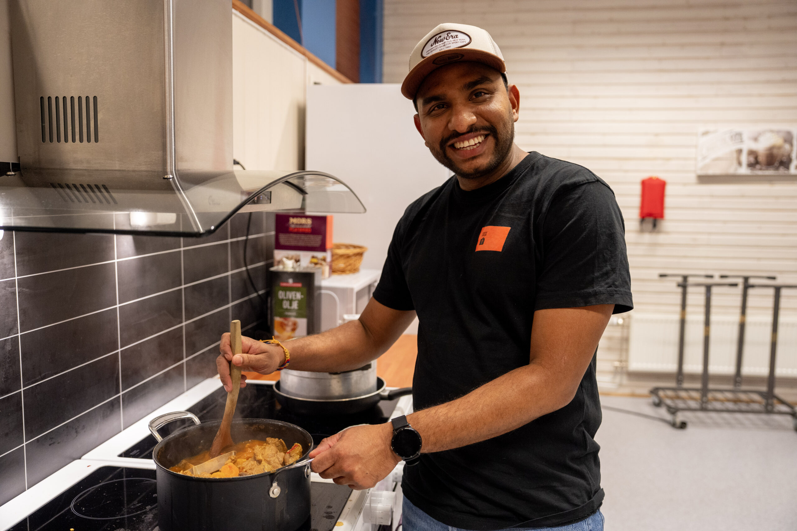 Smilende mann med caps som ser mot kamera. Han står over en kasserolle og rører.