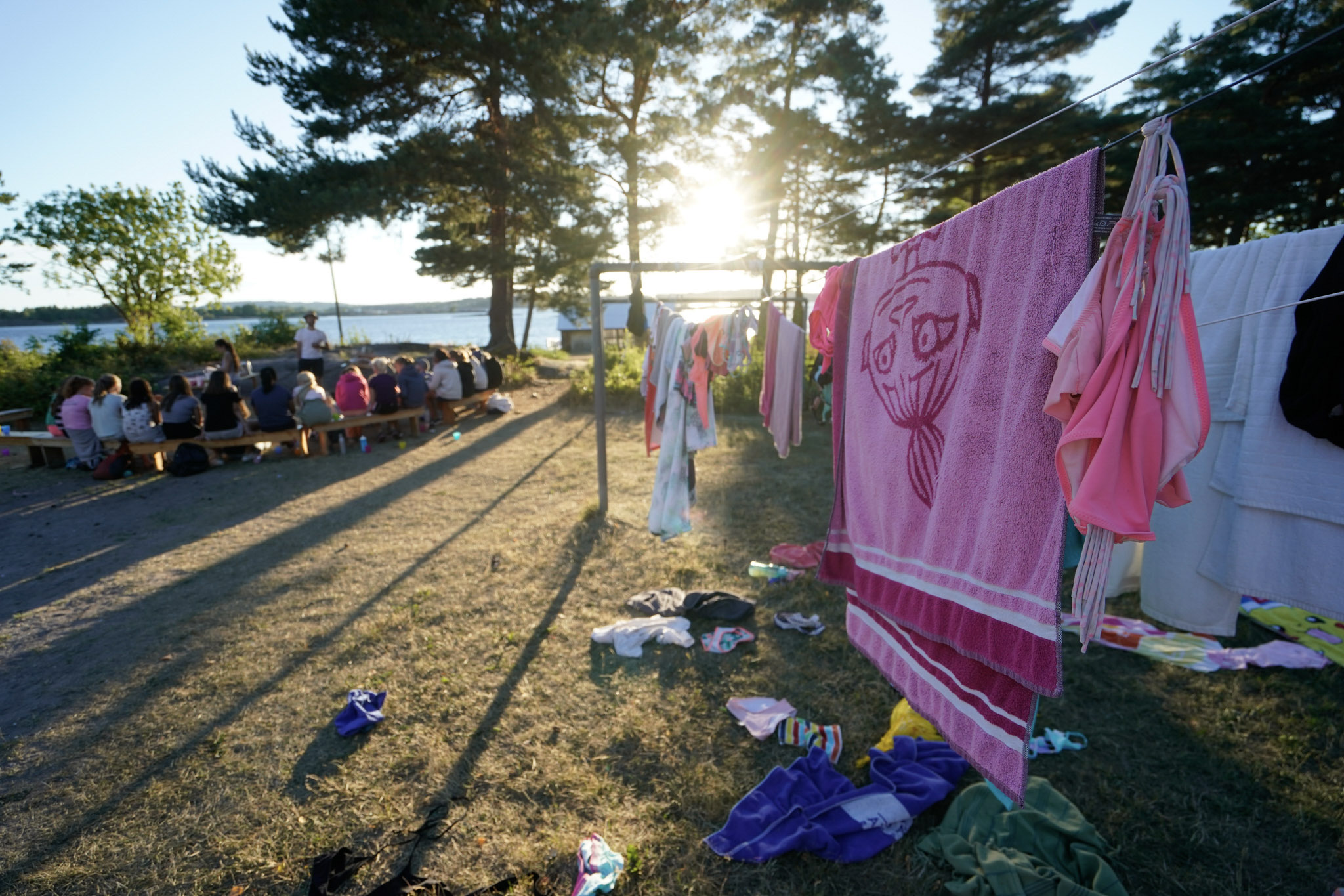 Håndklær henger til tørk utendørs i sola, en gjeng barn sitter samlet rundt et bord i bakgrunnen