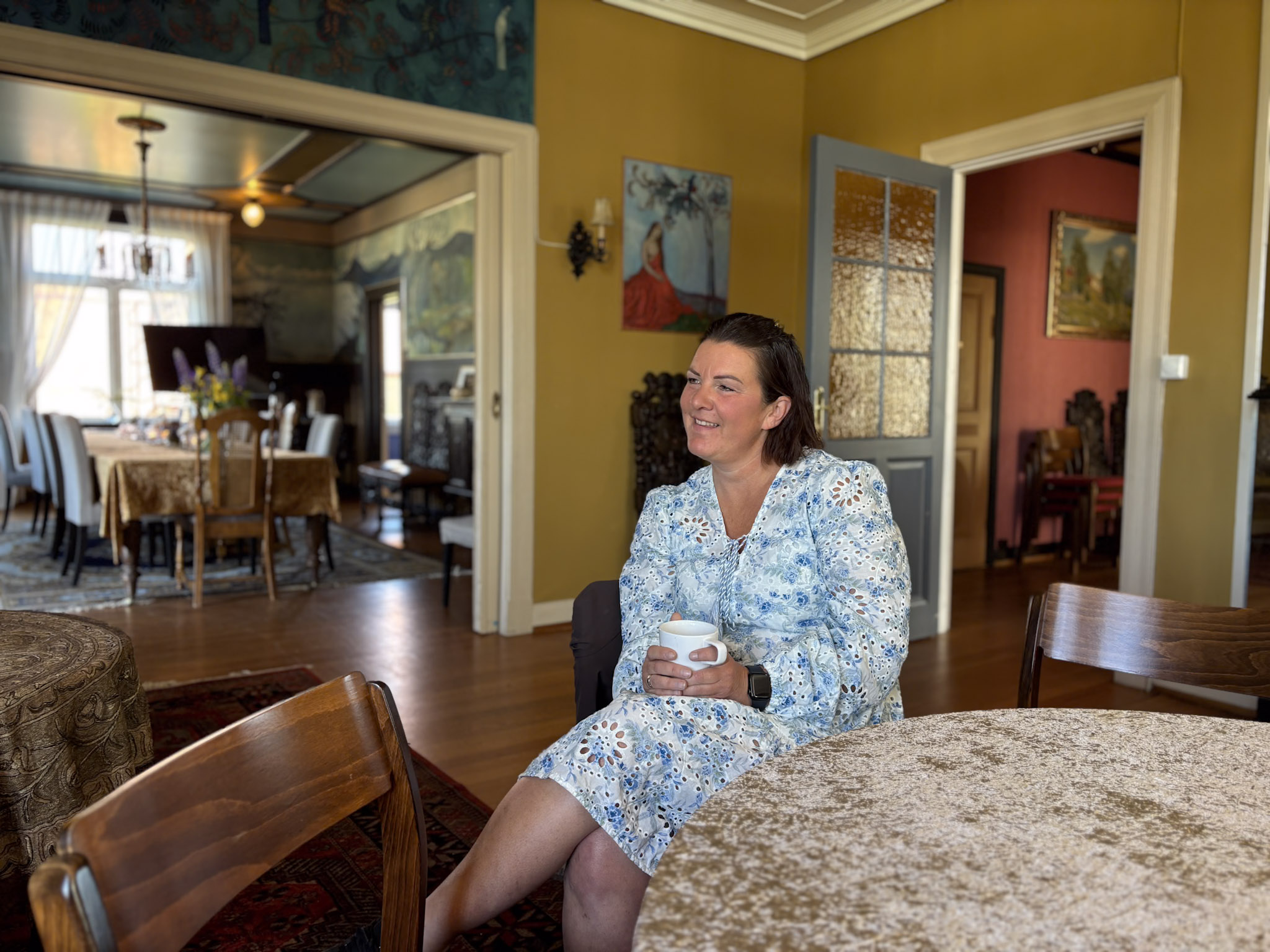Lene sitter ved et bord og smiler mens hun holder en kaffekopp.