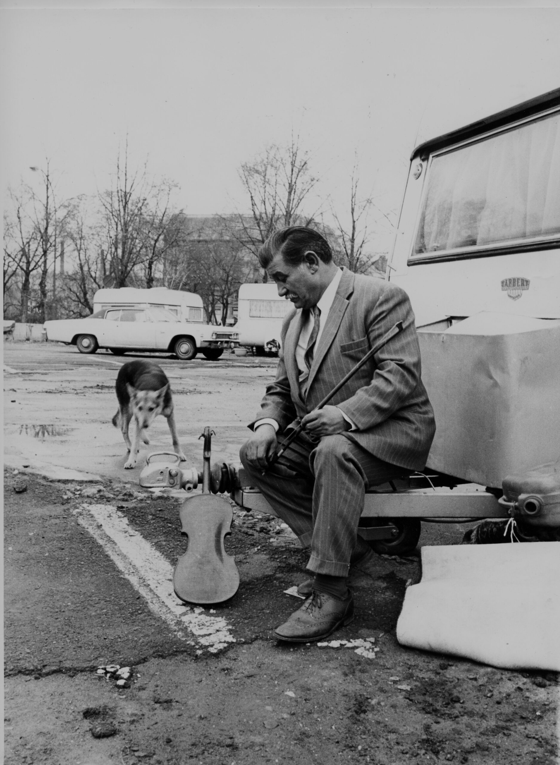 En mann utenfor en campingvogn på asfalt, han holder en fiolin, det er en hund i bakgrunnen.