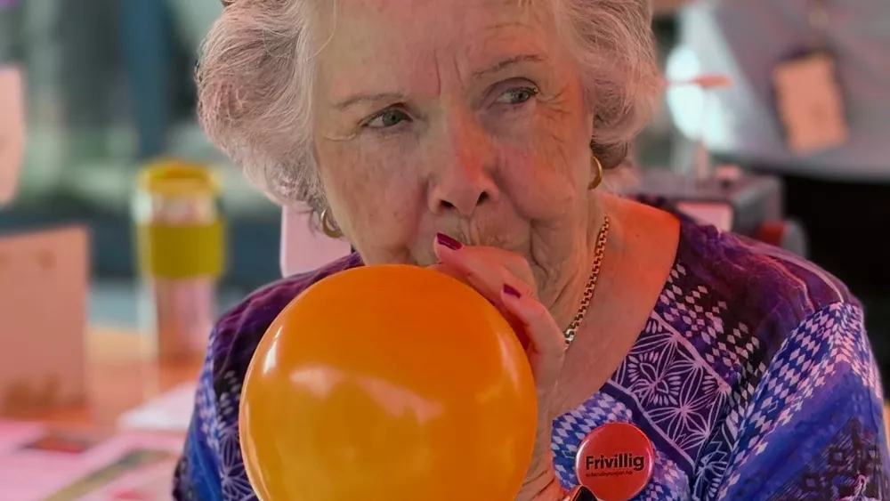 En eldre dame med frivillig-button bl&aring;ser opp en oransje ballong
